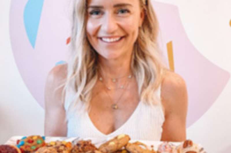 Vicki Melo smiles as she holds a tray of Sugar Mama Cookies in front of a pastel painting of a cookie with a bite taken out of it.