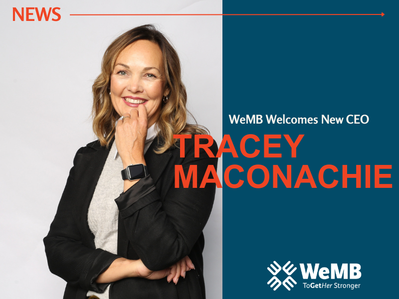 Tracey Maconachie, new CEO of WeMB, stands in front of a grey background wearing a black blazer over a grey shirt. She is smiling and has one hand up near her face.