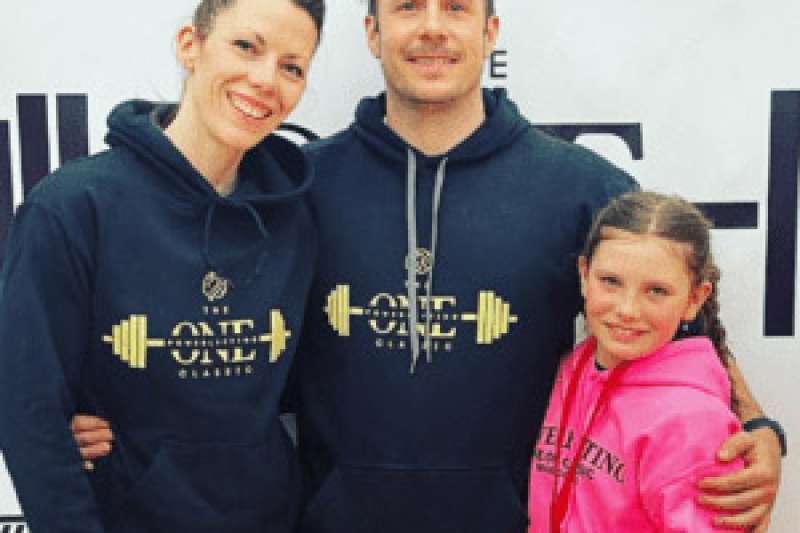 Kyla Lambert Camiré and her husband, Dino Camiré stand with their daughter. They are all wearing One Family Fitness hoodies.